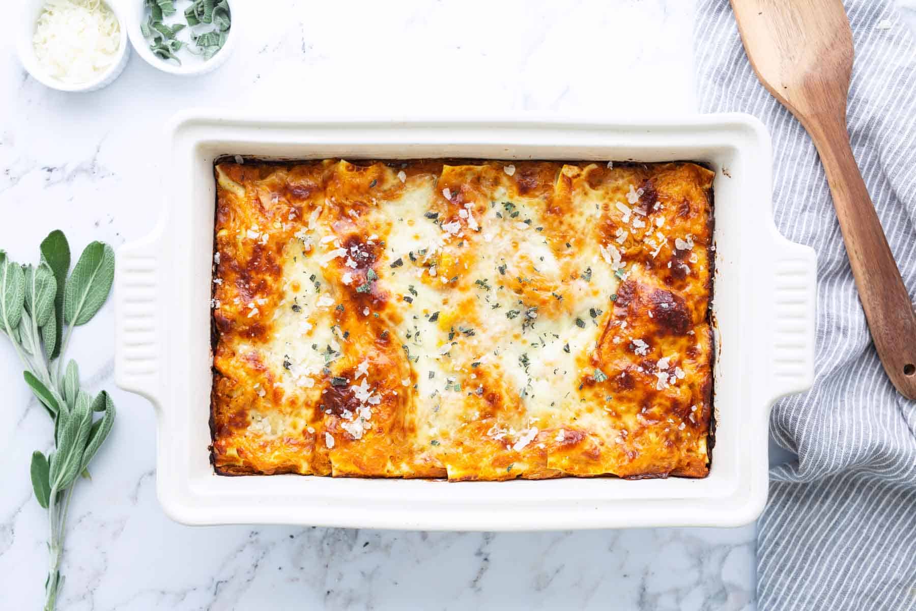 Baked lasagna in a white rectangular dish, topped with melted cheese and herbs, on a marble surface with a wooden spoon, herbs, and grated cheese nearby.