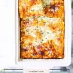 A baking dish with butternut squash lasagna, topped with melted cheese and herbs, with one square being served on a wooden spatula.