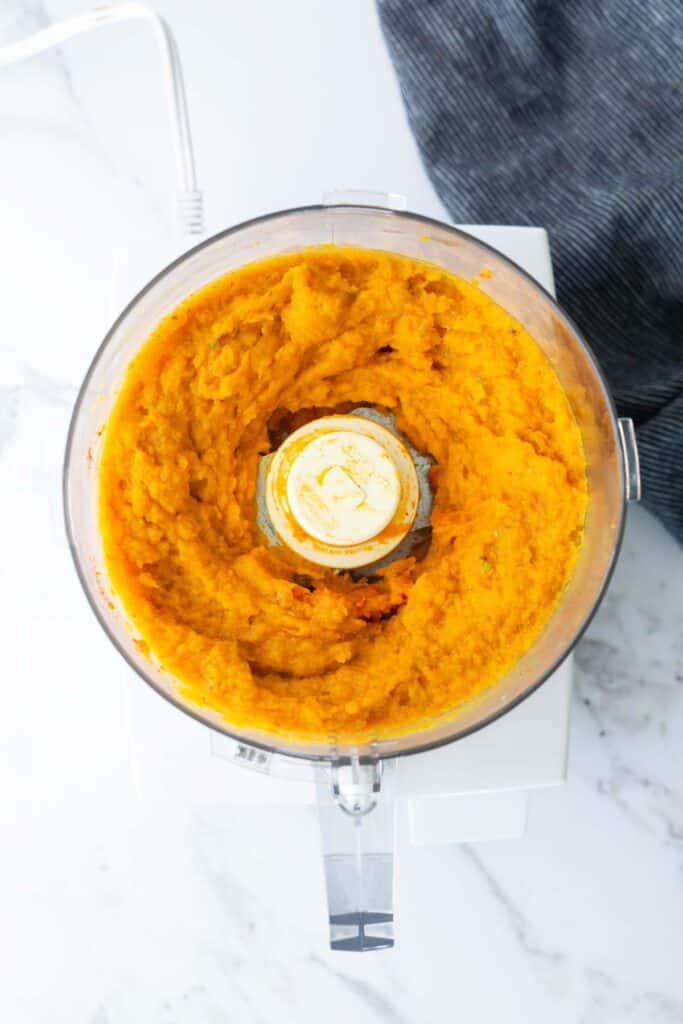 A food processor filled with smooth, blended orange pumpkin puree sits on a white countertop next to a dark blue cloth.