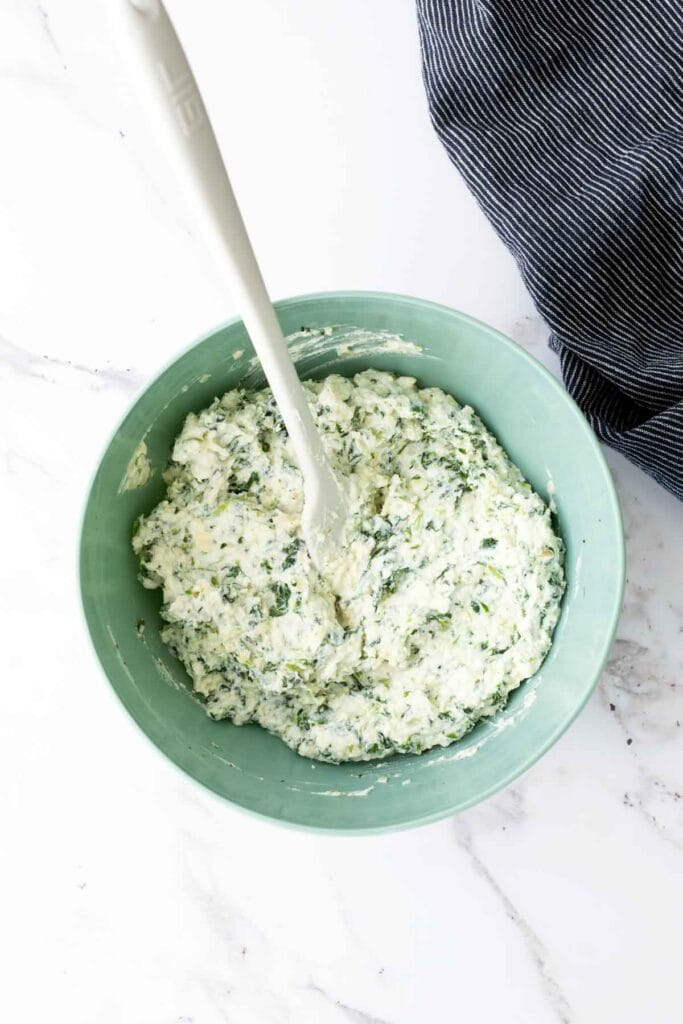 A green bowl filled with a creamy spinach and cheese mixture is shown with a white spatula, placed on a marble countertop next to a blue-striped cloth.