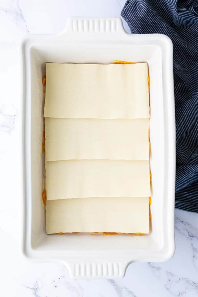Uncooked lasagna noodles arranged in a single layer in a white rectangular baking dish on a marble surface, next to a blue striped cloth.