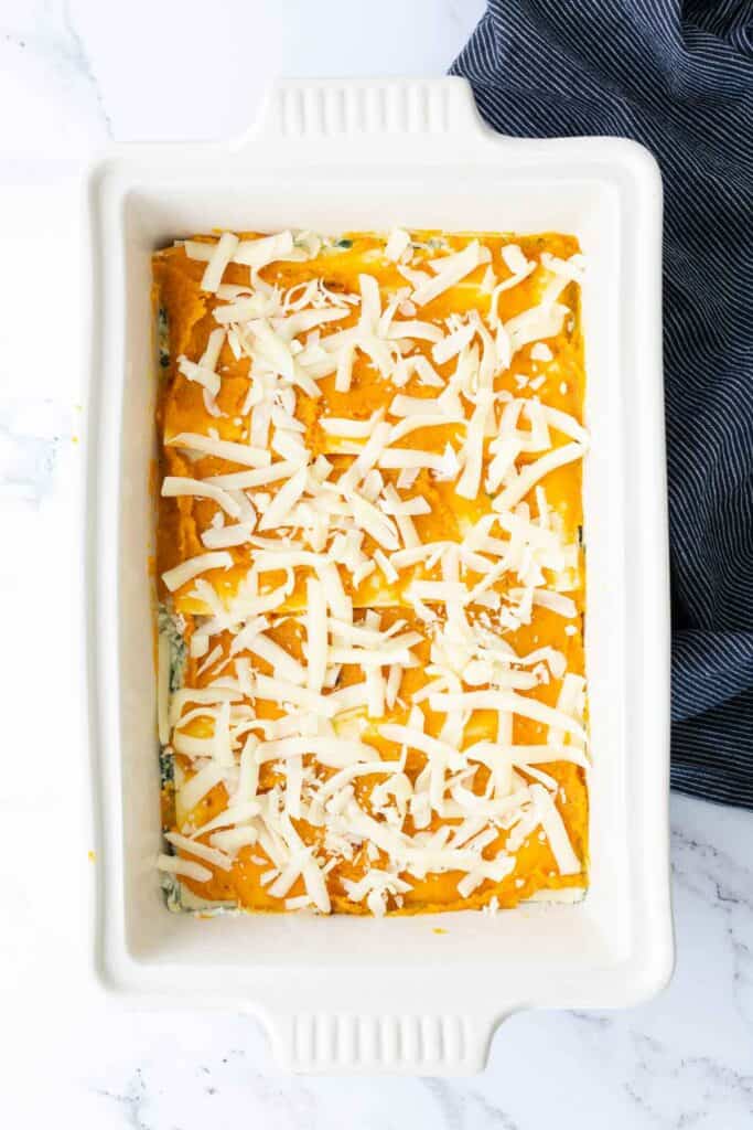 Rectangular white baking dish filled with a layered casserole, topped with shredded cheese, next to a dark blue striped cloth on a white marble surface.