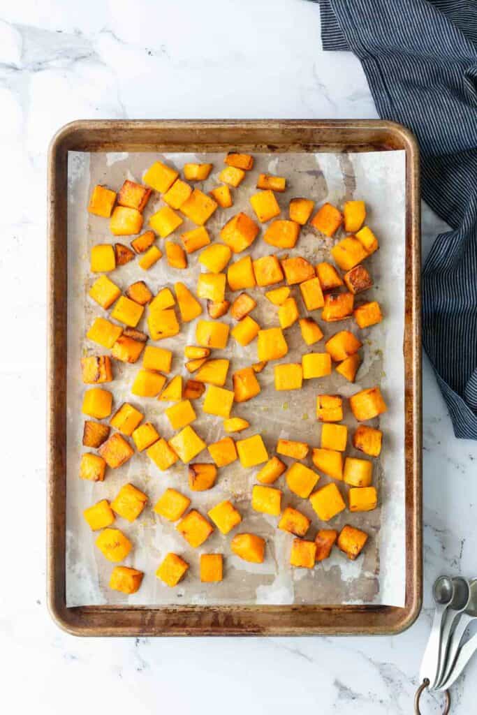 Baking sheet with evenly spaced roasted butternut squash cubes on parchment paper, next to a dark striped cloth and metal measuring spoons, on a marble countertop.