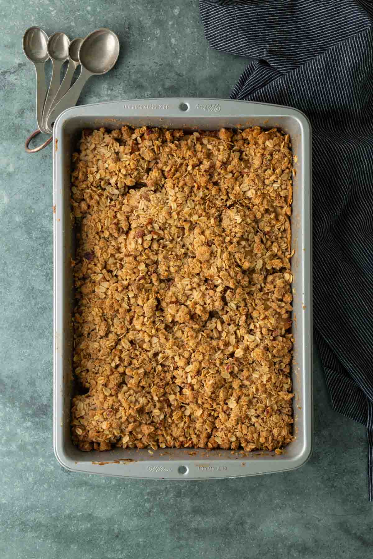 Rectangular baking dish filled with a golden-brown oat crumble sits on a green countertop next to a black-striped cloth and three nested measuring spoons.