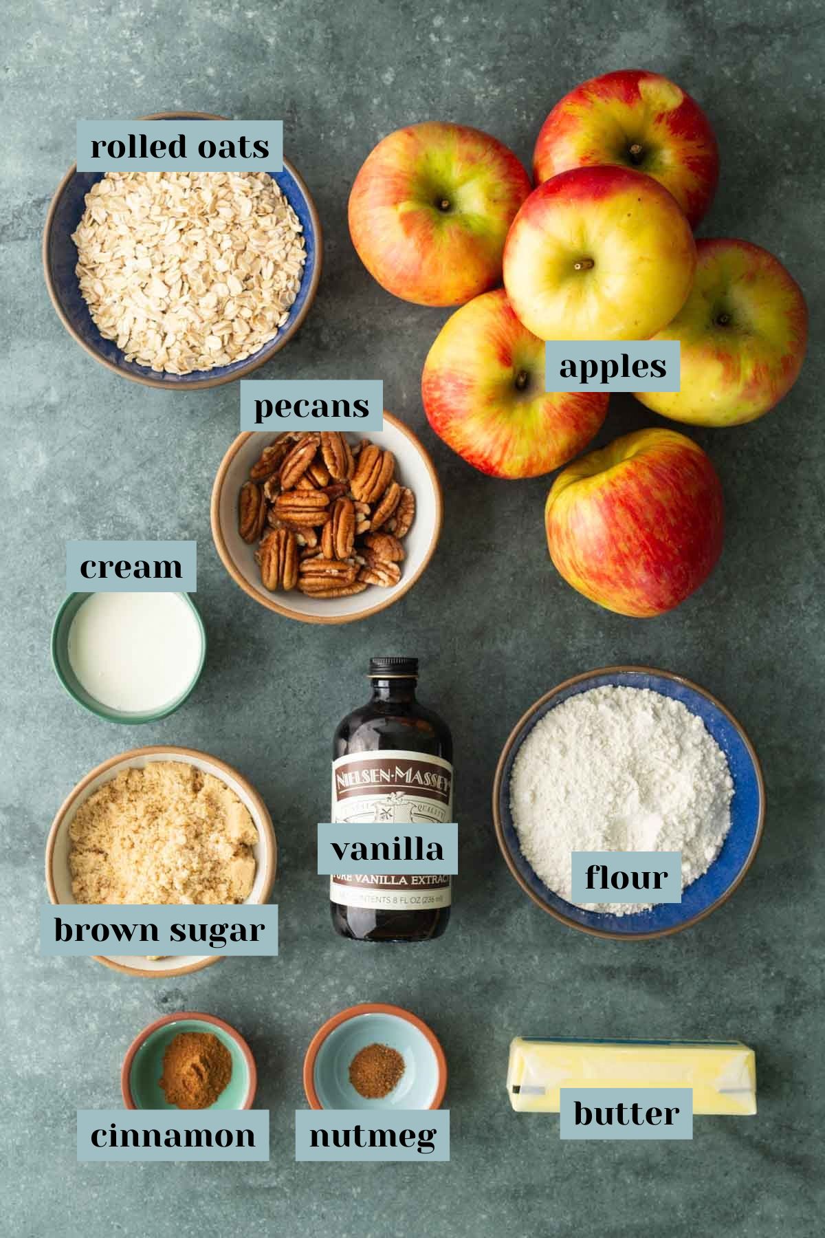 A flat lay of ingredients for baking: rolled oats, apples, pecans, cream, flour, brown sugar, vanilla extract, cinnamon, nutmeg, and butter, each labeled.