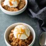 Two white bowls of apple crisp topped with vanilla ice cream and caramel sauce sit on a gray surface next to a spoon and a striped cloth.