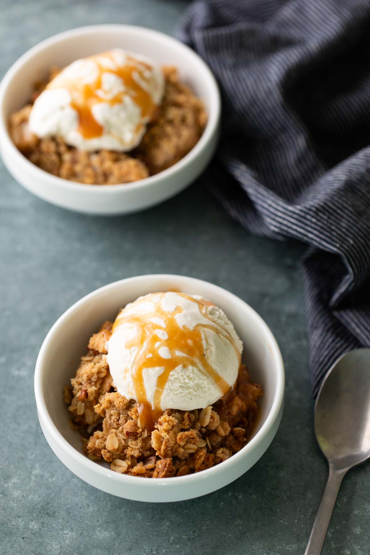 Two white bowls of apple crisp topped with vanilla ice cream and caramel sauce sit on a gray surface next to a spoon and a striped cloth.