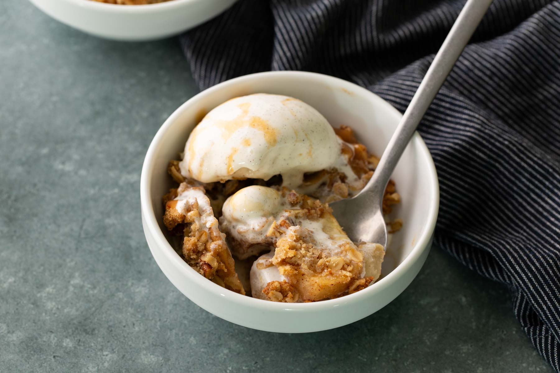 A bowl of apple crisp topped with a scoop of melting vanilla ice cream, with a spoon resting inside the bowl.