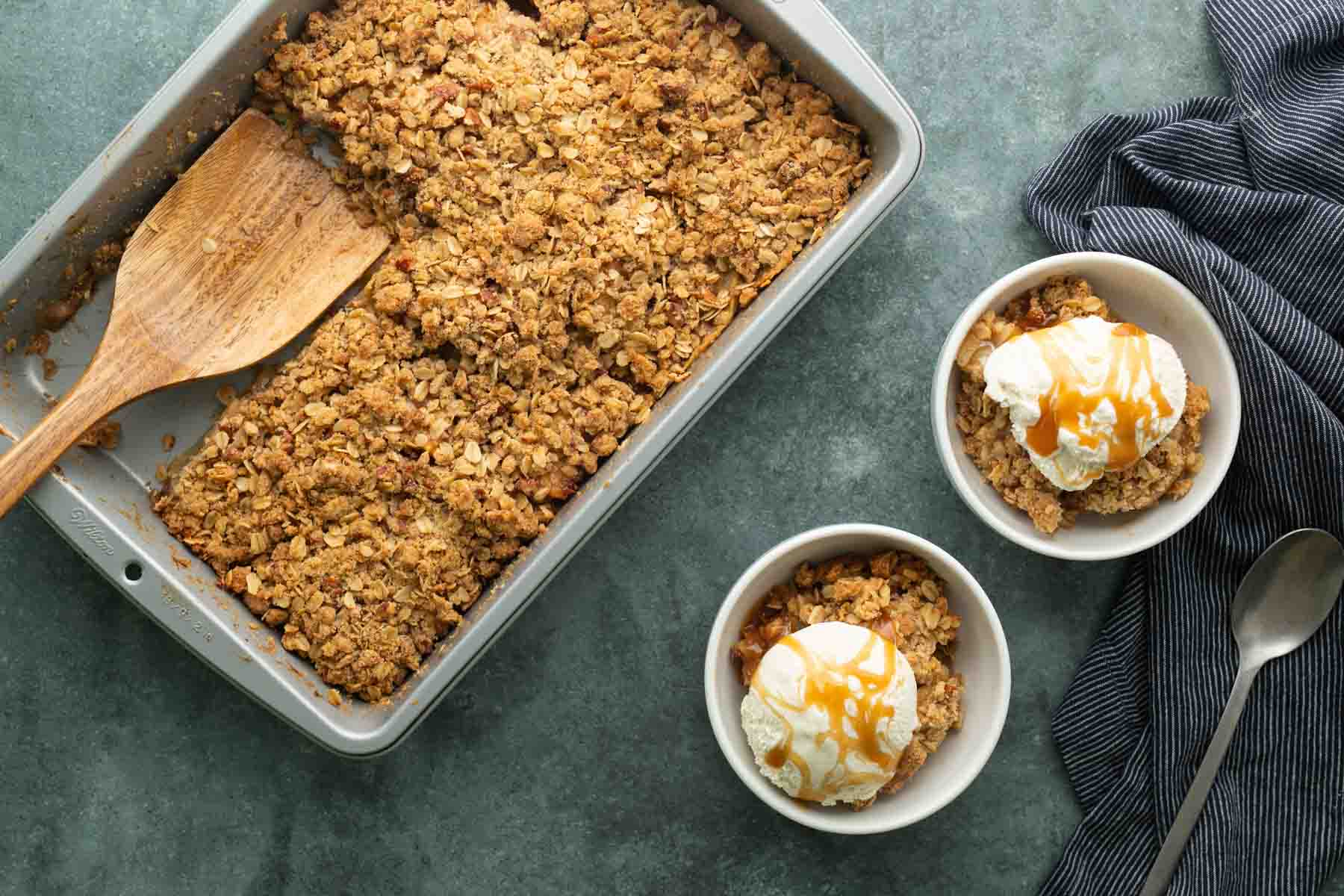 A baking pan with fruit crisp and a wooden spatula, next to two bowls of crisp topped with vanilla ice cream and caramel sauce, a spoon, and a striped cloth.