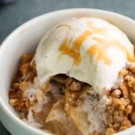A bowl of caramel apple crisp topped with melting vanilla ice cream and drizzled with caramel sauce.