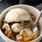 A bowl of caramel apple crisp topped with a scoop of vanilla ice cream, with a spoon inside the bowl.