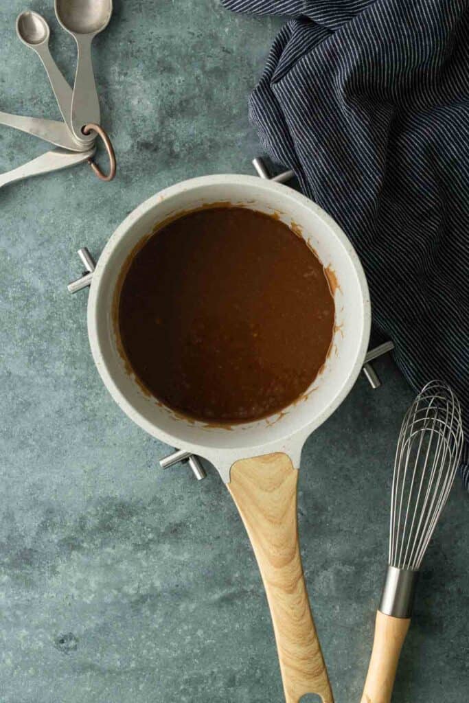 A saucepan filled with brown sauce sits on a blue-gray countertop, surrounded by a whisk, measuring spoons, and a striped cloth.