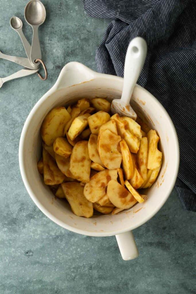 White mixing bowl with sliced apples coated in spices, a white spoon inside, metal measuring spoons, and a striped cloth on a green countertop.