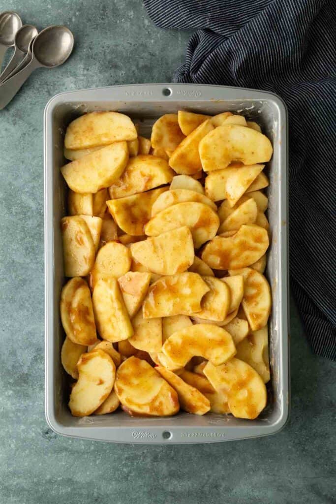 Rectangular baking dish filled with sliced apples coated in a light brown mixture, set on a green countertop next to a blue-striped cloth and metal measuring spoons.