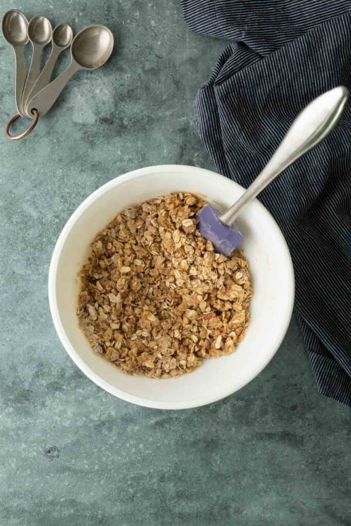 White bowl with oat crumble mixture and a spatula on a green countertop, with measuring spoons and a striped cloth nearby.