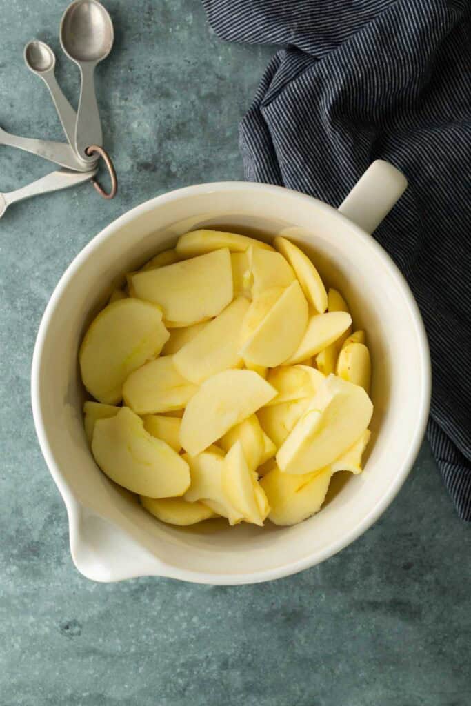A white mixing bowl filled with sliced apples sits on a blue countertop next to metal measuring spoons and a dark striped cloth.