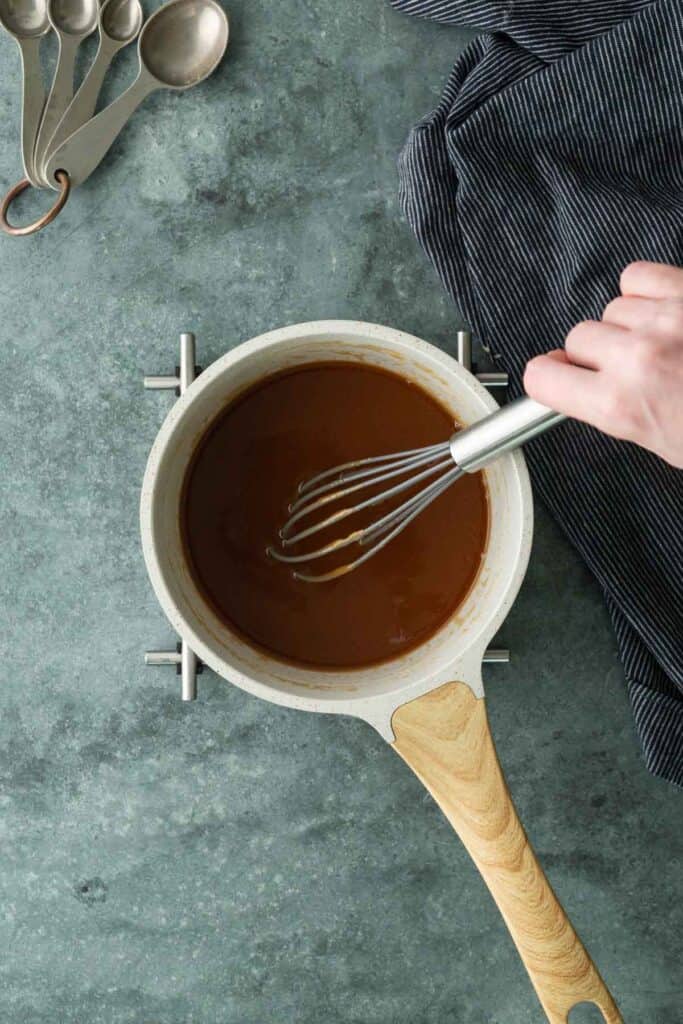 Hand whisking brown sauce in a white saucepan on a stovetop, with metal measuring spoons and a striped cloth nearby on a gray countertop.