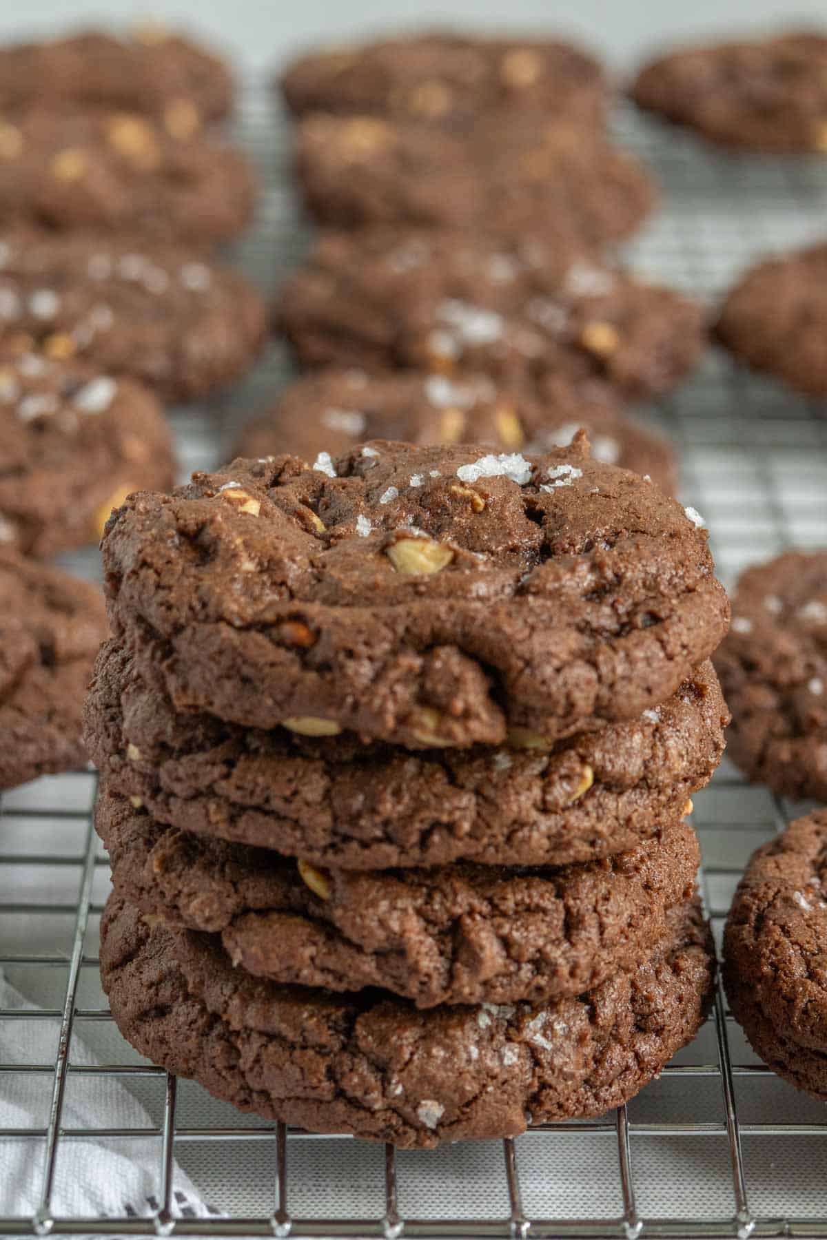 A stack of chocolate cookies with sea salt and nuts sits on a wire cooling rack, with more cookies cooling in the background.