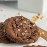 Two chocolate cookies with nuts and flaky salt rest on parchment paper, with crumbs and a blurred milk container in the background.