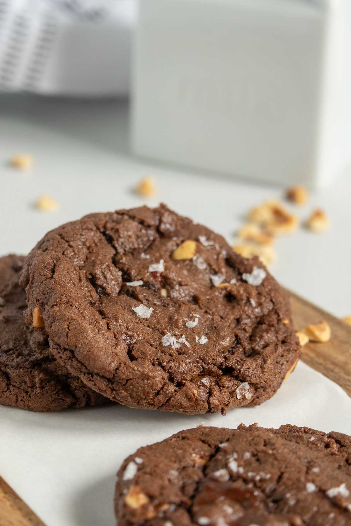 Two chocolate cookies with nuts and flaky salt rest on parchment paper, with crumbs and a blurred milk container in the background.