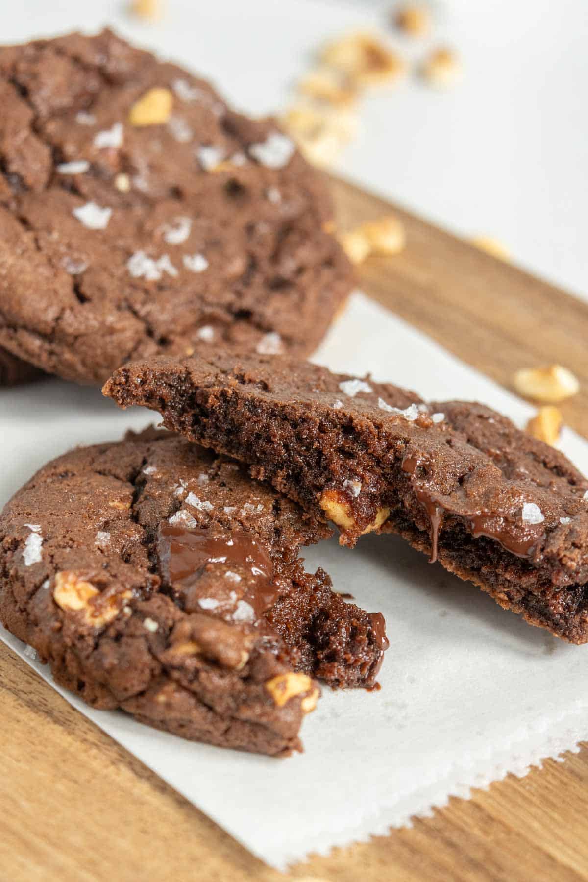 Two chocolate cookies with nuts are on parchment paper, one broken in half to show a gooey chocolate center. A few cookie crumbs and nuts are scattered around on a wooden surface.