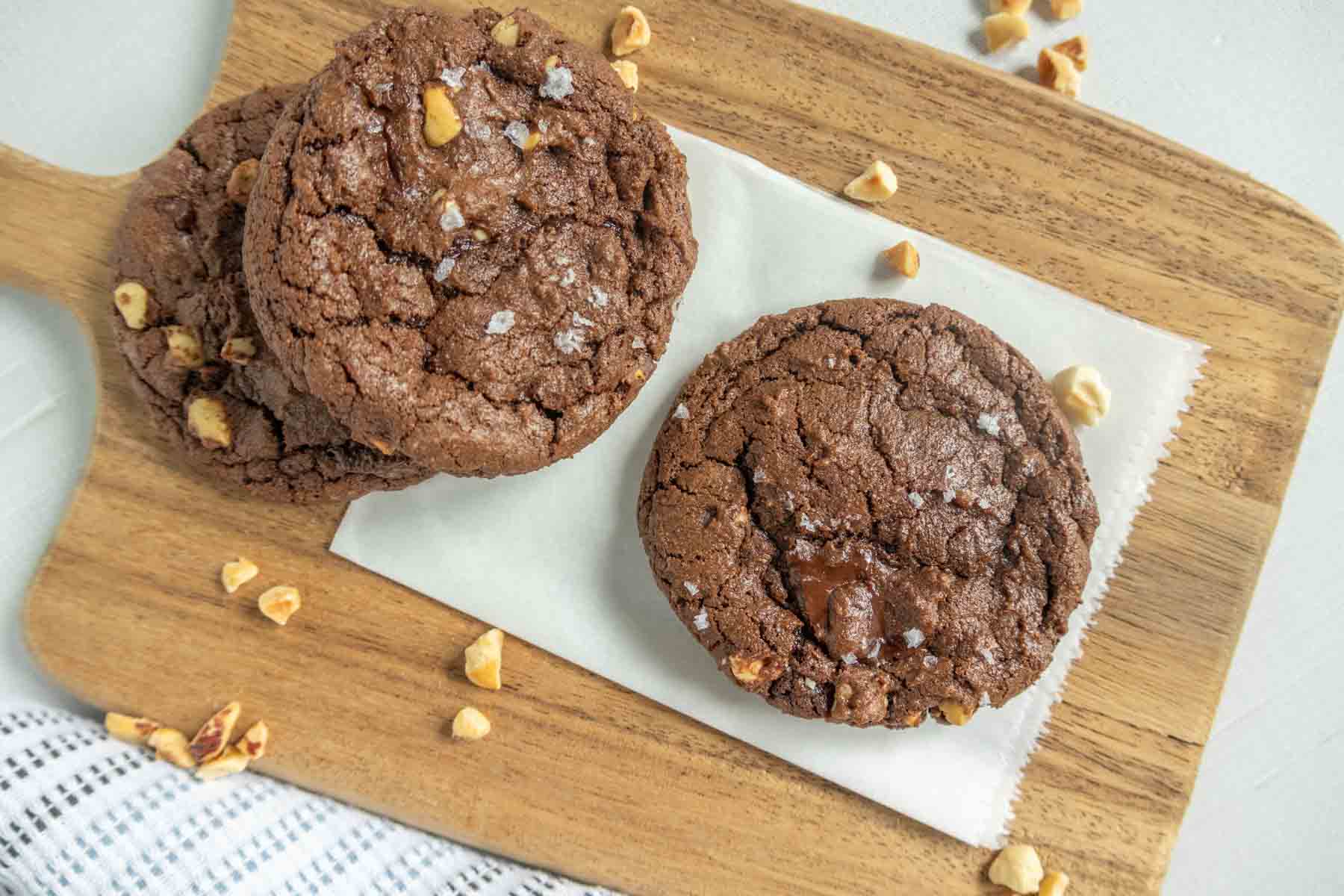 Two chocolate cookies with nuts sprinkled on top, placed on a piece of parchment paper on a wooden serving board, with extra nut pieces scattered around.