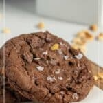 Two chocolate hazelnut cookies topped with flaky salt are stacked on a surface, with scattered hazelnut pieces and a blurred kitchen background.