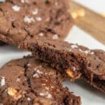 Close-up of chocolate hazelnut cookies on parchment paper, one broken in half showing melted chocolate and hazelnut pieces, topped with flaky salt.