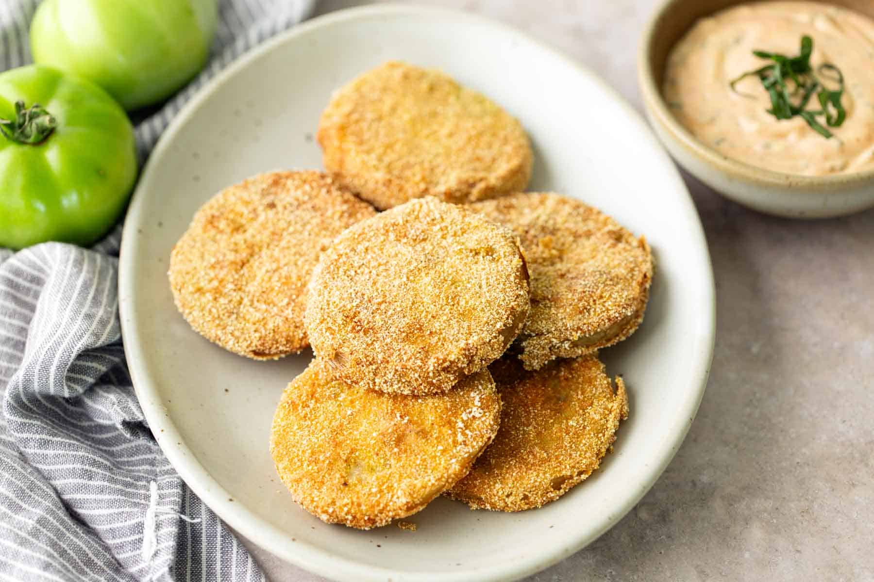A plate of breaded fried green tomato slices with a bowl of dipping sauce and two whole green tomatoes in the background.