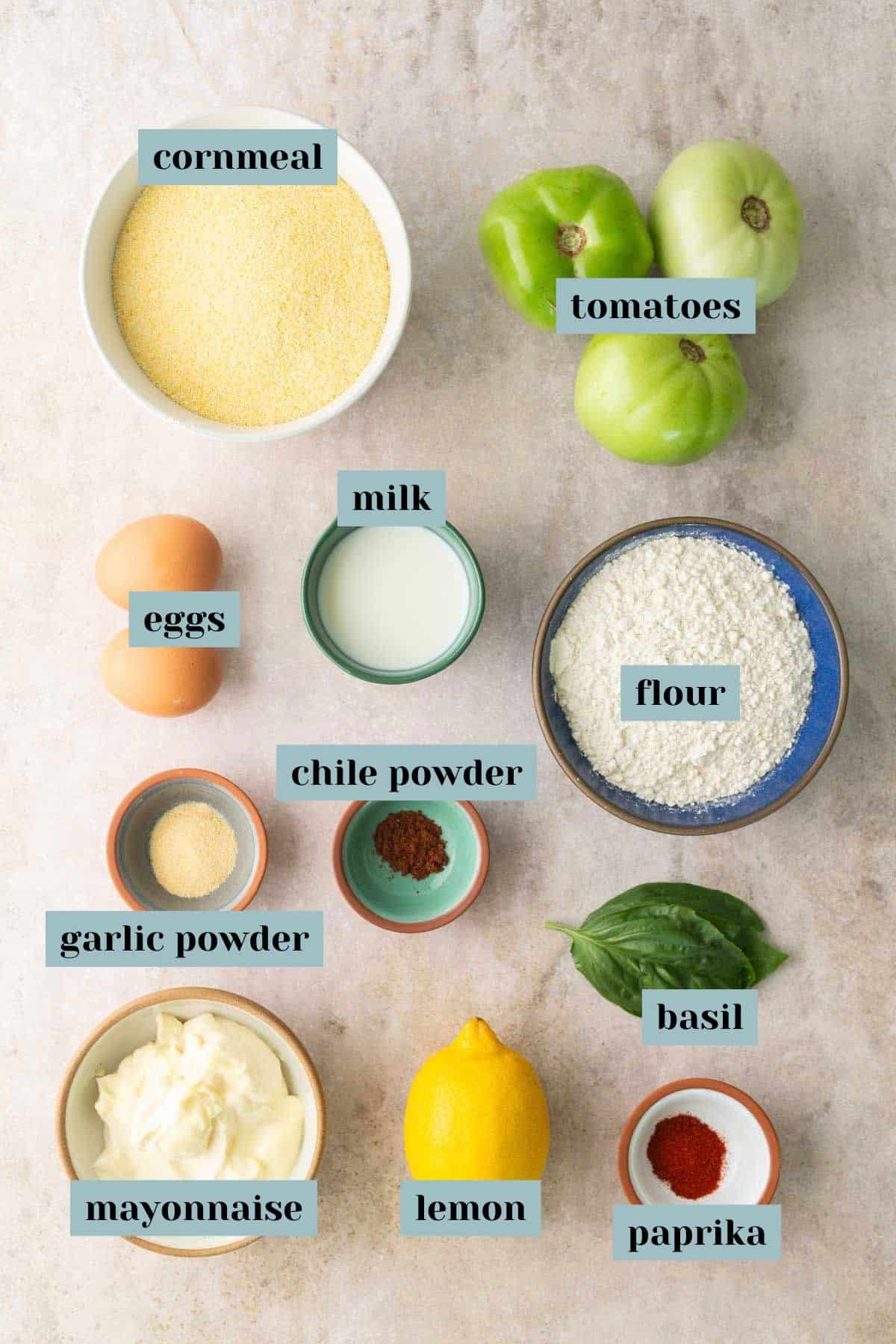 Assorted ingredients including cornmeal, tomatoes, eggs, milk, flour, mayonnaise, lemon, basil, and spices arranged on a white background.