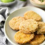 A plate of breaded and fried green tomato slices sits on a white dish, with a bowl of dipping sauce and two whole green tomatoes in the background.
