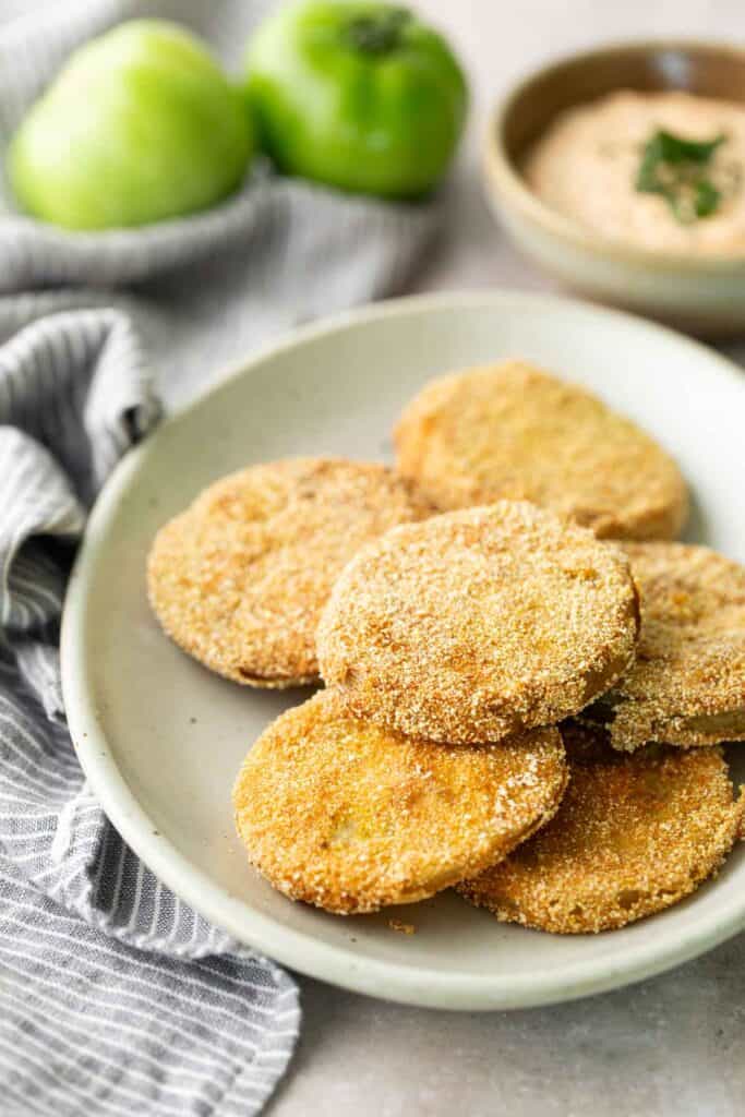 Fried Green Tomatoes A plate of breaded and fried green tomato slices sits on a white dish, with a bowl of dipping sauce and two whole green tomatoes in the background.
