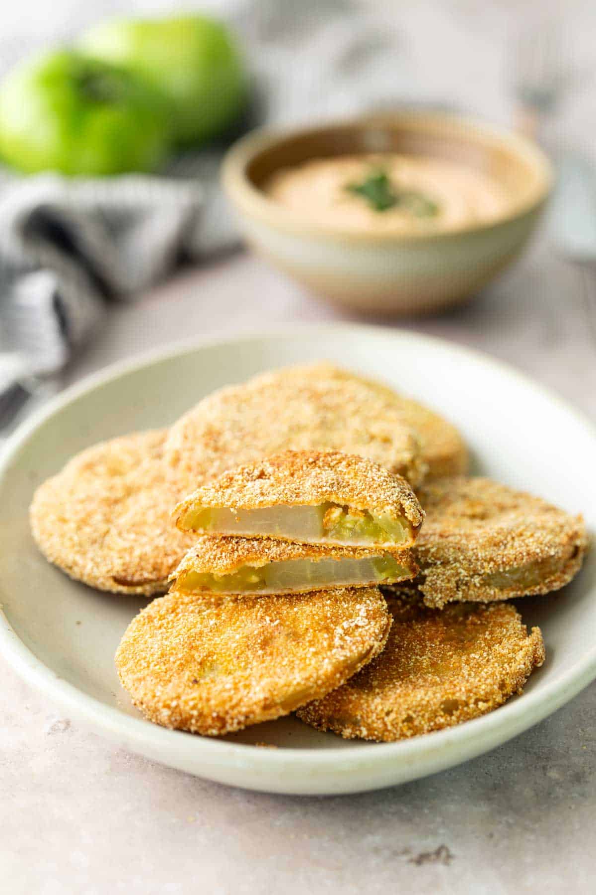 Plate of golden-brown fried green tomato slices with a dipping sauce in the background, one slice cut in half to show the inside.