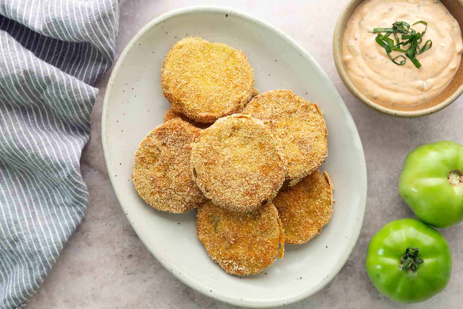 A plate of fried green tomato slices coated in cornmeal sits on a table next to a bowl of dipping sauce and two raw green tomatoes.