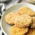 A plate of breaded and fried green tomato slices sits on a table, with green tomatoes and a bowl of dipping sauce in the background.