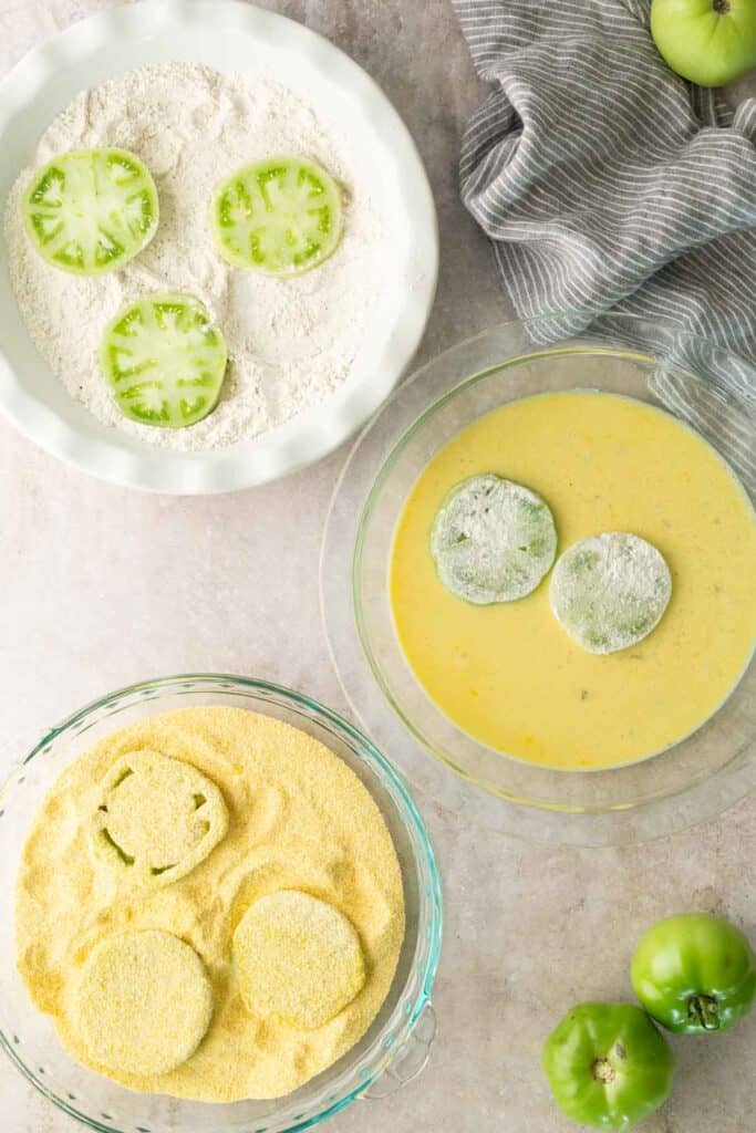 Three green tomato slices being coated in flour, egg mixture, and cornmeal, shown step-by-step in separate bowls on a countertop with a striped towel and whole green tomatoes nearby.