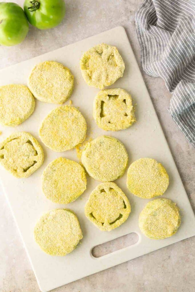 Slices of green tomatoes coated in cornmeal sit on a white cutting board next to a gray striped towel and whole green tomatoes.