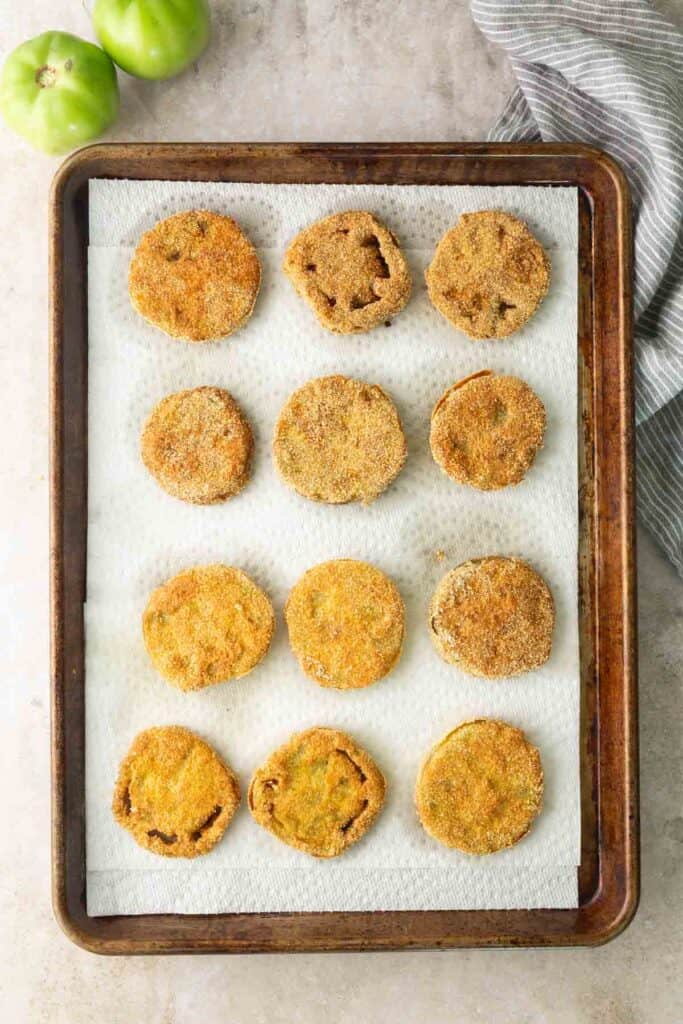 Twelve breaded and fried green tomato slices arranged in rows on a parchment-lined baking sheet, with two whole green tomatoes and a striped cloth nearby.