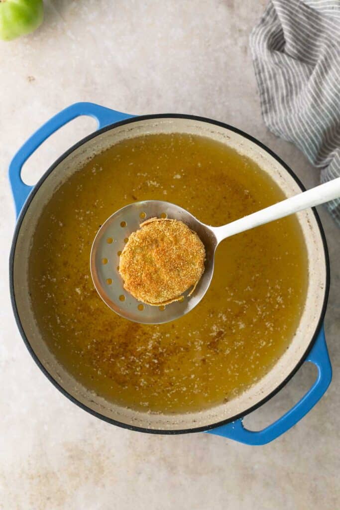 A breaded round slice is held on a slotted spoon above a pot of hot oil, ready for frying. A striped towel is visible in the top right corner.