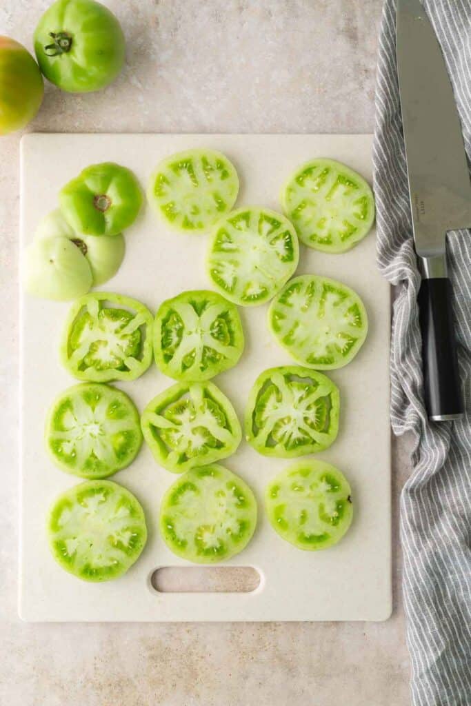 Sliced green tomatoes are arranged on a white cutting board next to a chef’s knife and a gray striped towel.