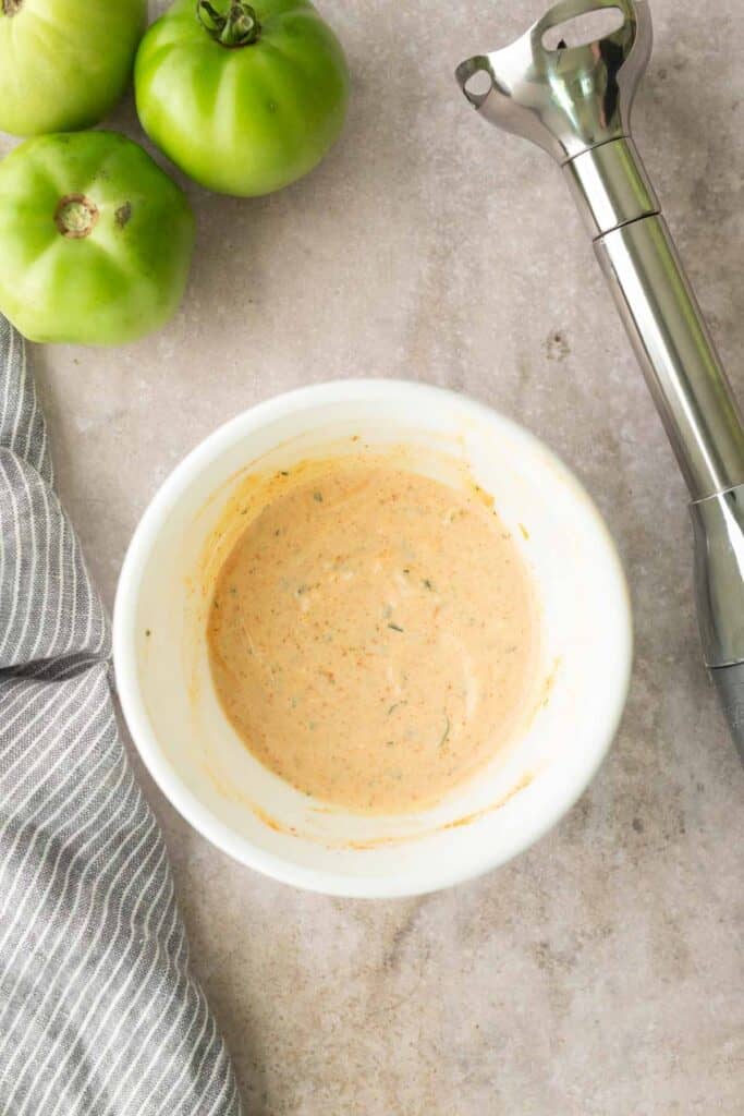 A white bowl of creamy sauce on a gray countertop, next to green tomatoes, a striped cloth, and an immersion blender.