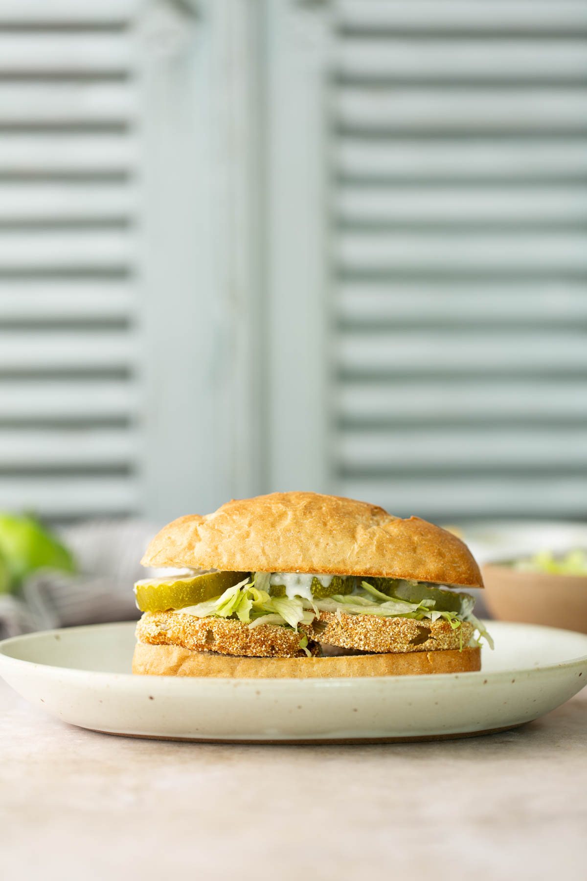 A fried green tomato sandwich with lettuce, pickles, and sauce is served on a round plate in front of a light blue shuttered background.