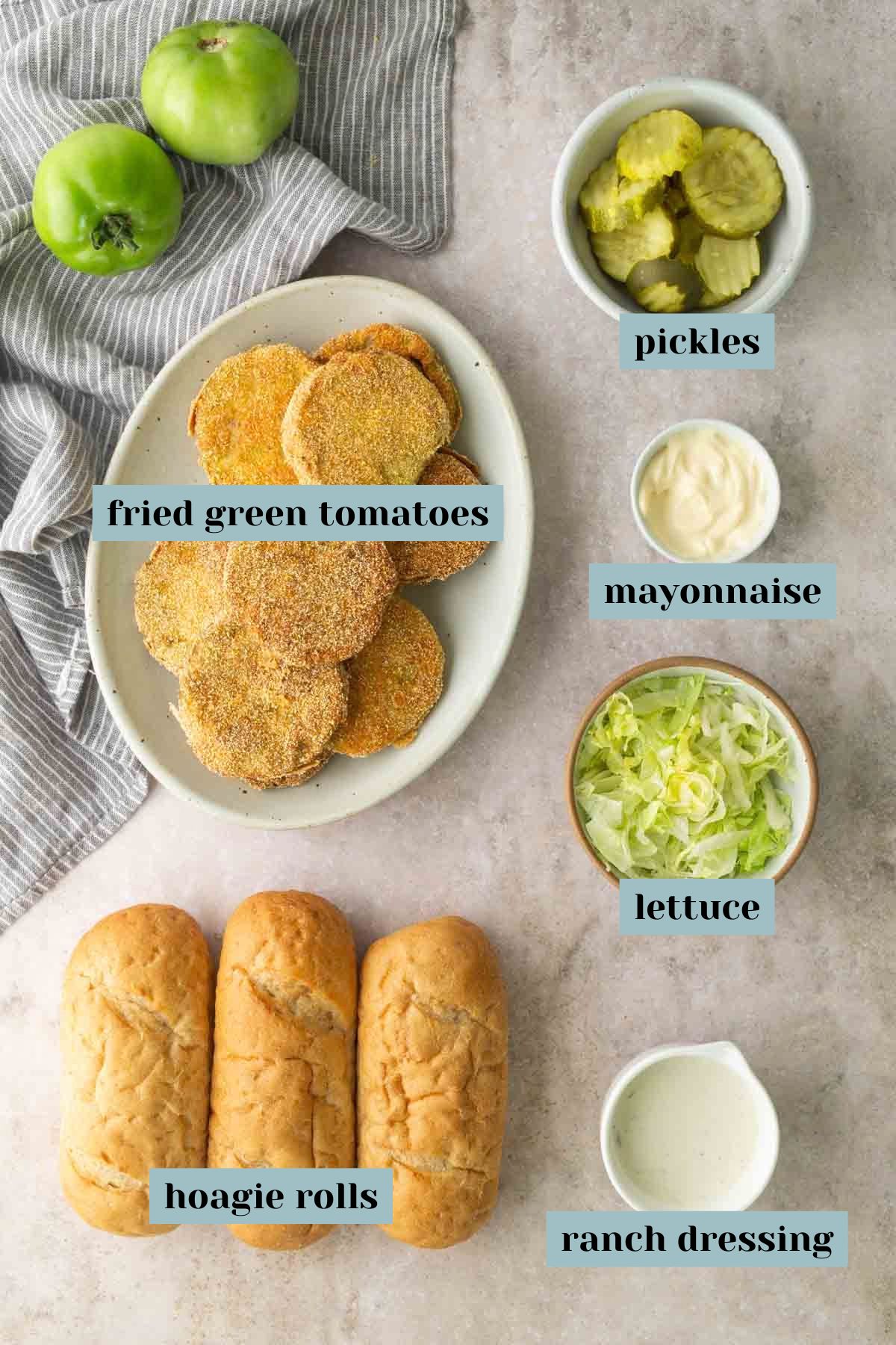 A plate of fried green tomatoes, pickles, mayonnaise, shredded lettuce, three hoagie rolls, and ranch dressing are arranged on a countertop.