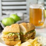 A sandwich with lettuce and fried green tomatoes on a plate with potato chips and coleslaw; a glass of beer and whole green tomatoes are in the background.
