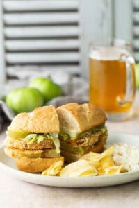 A sandwich with lettuce and fried green tomatoes on a plate with potato chips and coleslaw; a glass of beer and whole green tomatoes are in the background.
