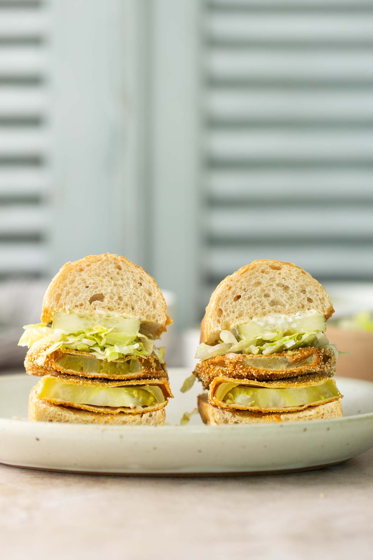 Two halves of a sandwich with layered fried vegetables, lettuce, and sliced bread are displayed on a plate in front of a blurred background.