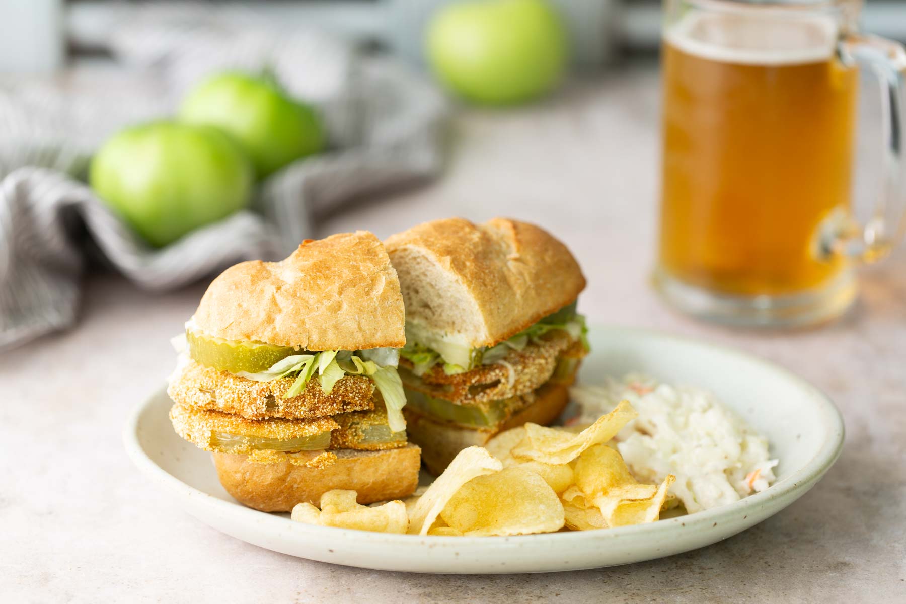 A plate with a fried green tomato sandwich, potato chips, and coleslaw. A glass of beer and green tomatoes are in the background.