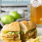 A fried green tomato po boy sandwich with lettuce on a baguette is served with potato chips on a plate; green tomatoes and iced tea are in the background.