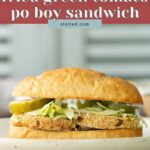A fried green tomato po’ boy sandwich with lettuce, pickles, and sauce on a bun, served on a plate.