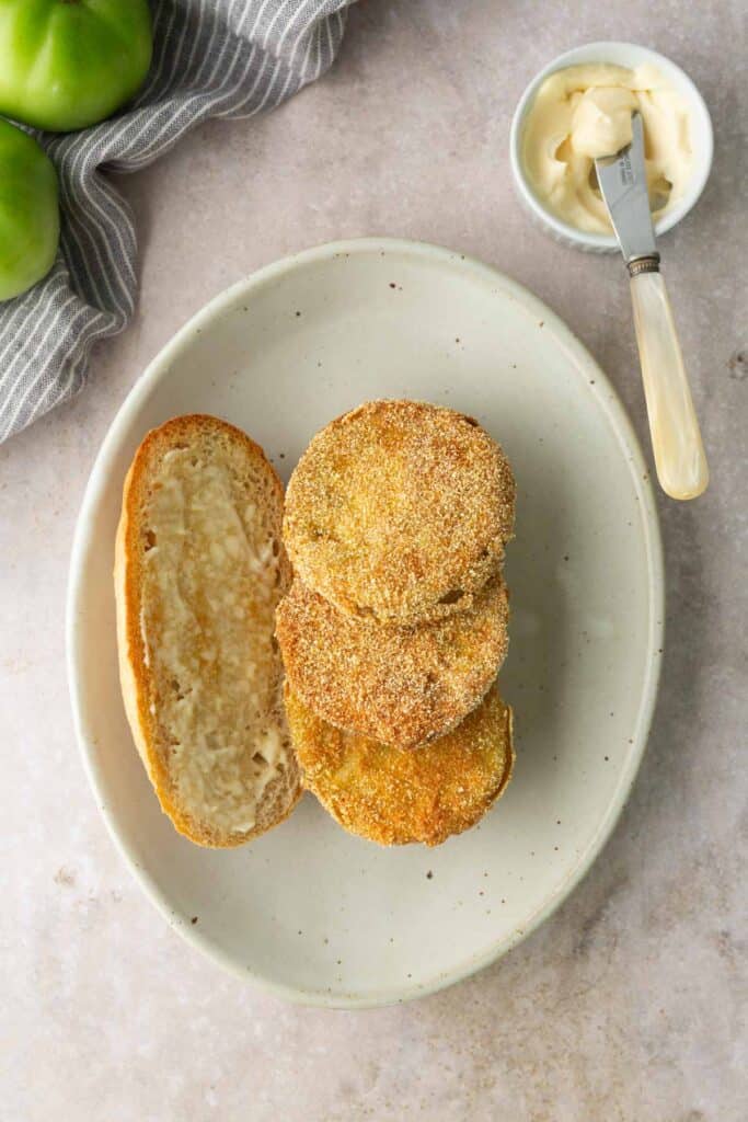 Three fried green tomato slices on an open sandwich bun, served on an oval plate with a side of mayonnaise and a spreading knife. Green tomatoes and a striped cloth are in the background.